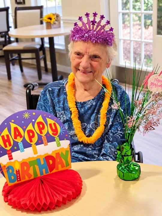 A resident in a purple tiara and yellow lei beside a Happy Birthday centerpiece