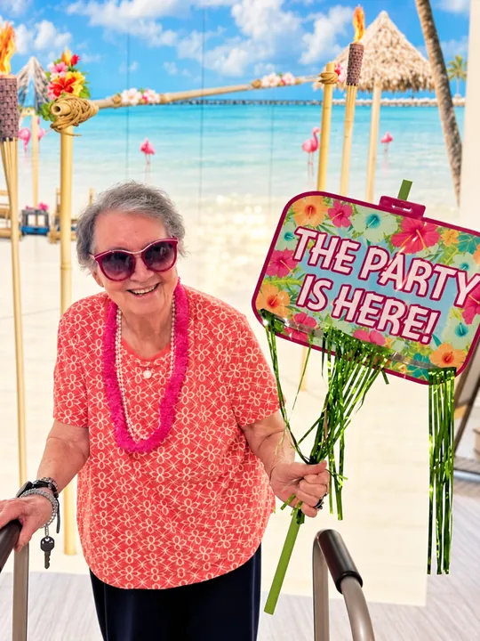 A resident wearing a lei and sunglasses in front of a beach backdrop, holding a sign that reads The Party Is Here