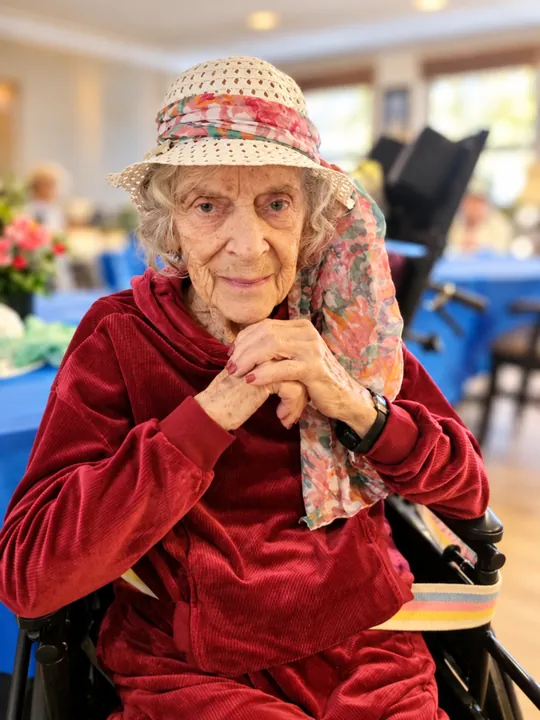 A resident in a red velvet jacket and straw hat seated in the dining room