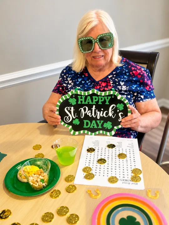 A resident in shamrock glasses holding a Happy St. Patrick's Day sign at a table with gold coins and a word search